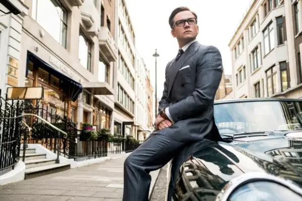 Taron Egerton in Kingsman: The Golden Circle wearing a grey suit, sitting on a classic car