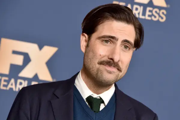 Jason Schwartzman wearing a black suit jacket and blue cardigan