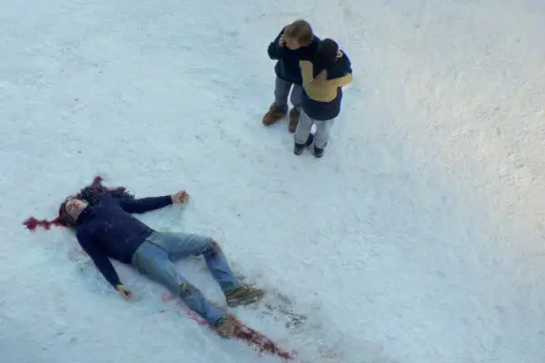 A man laying in the snow, dead, as two people stand next to him