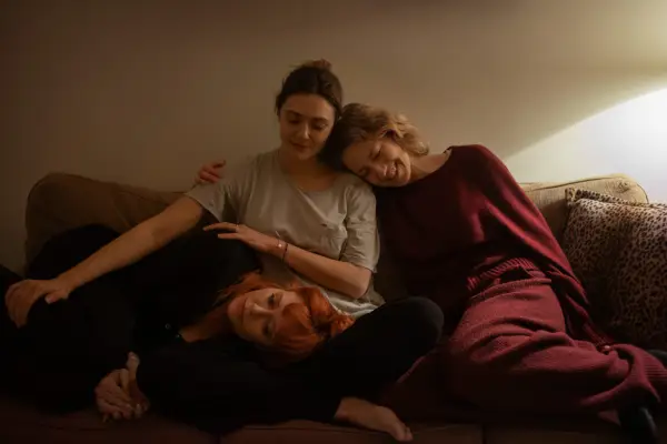 Natasha Lyonne as Rachel, Elizabeth Olsen as Christina and Carrie Coon as Katie in His Three Daughters sitting together in an embrace on a sofa and smiling.