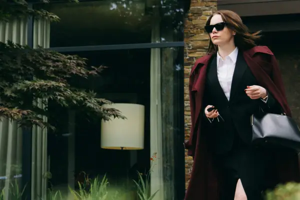 Emma Stone as Michelle Fuller in Bugonia, wearing a red velvet suit and sunglasses, walking out of a building.