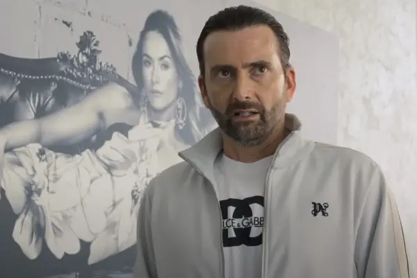 David Tennant in The Thursday Murder Club, wearing a white T shirt, grey jacket and snarling, with a picture of a woman on the wall behind him.