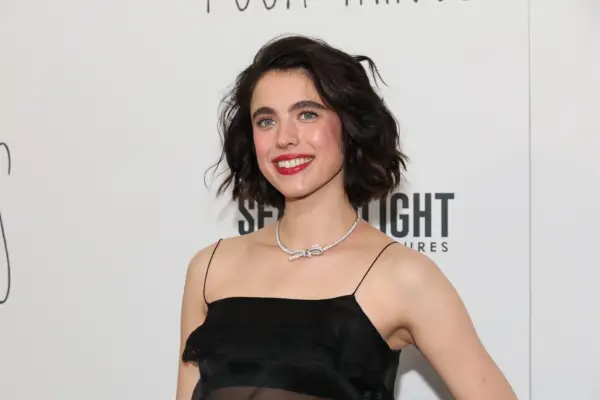 Margaret Qualley wearing a black dress and smiling, stood in front of a white background