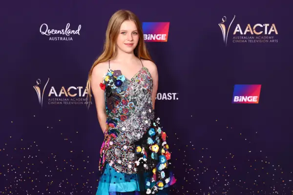 Alyla Browne wearing a shiny, flowery and blue dress and posing in front of a board bearing the name and logo of the Australia Academy Cinema Television Arts.