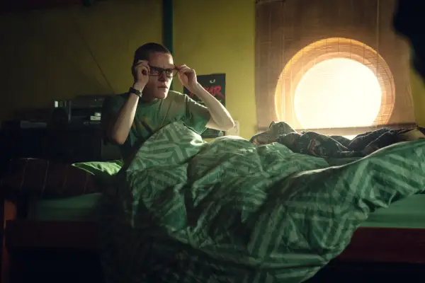 Lewis Gribben in Black Mirror season 7 holding his glasses to his eyes and waking up in a dimly lit room.