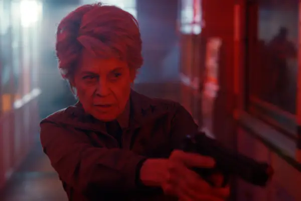 Linda Hamilton as Dr. Kay in Stranger Things illuminated by a red light as she walks through the dark hallways of a building holding a gun.