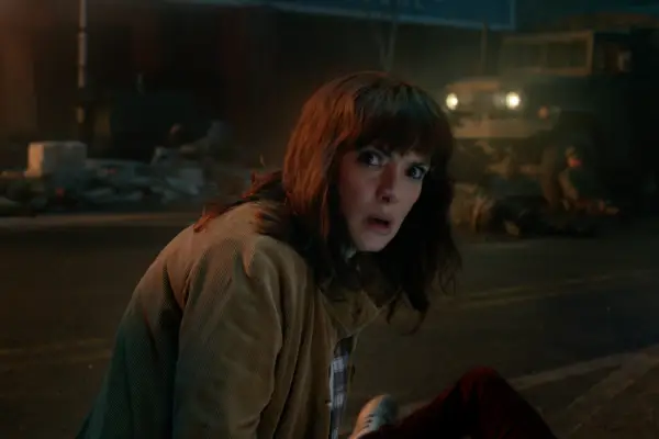 Winona Ryder as Joyce Byers in Stranger Things crouching down on a roadside and looking surprised as she looks to the side.