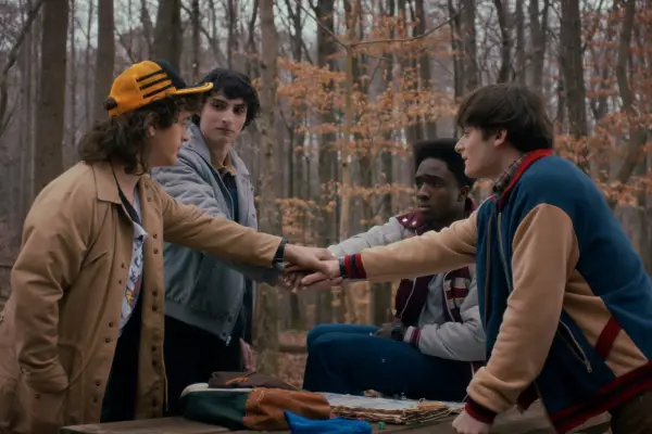 Gaten Matarazzo as Dustin Henderson, Finn Wolfhard as Mike Wheeler, Caleb McLaughlin as Lucas Sinclair, and Noah Schnapp as Will Byers in Stranger Things all putting their hands in the middle of the group and looking at each other while standing in the woods.