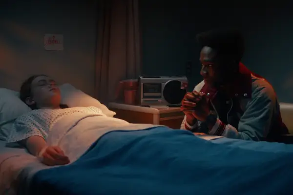 Sadie Sink as Max Mayfield in a hospital bed with her eyes closed while Caleb McLaughlin sits by her bed and looks worried.