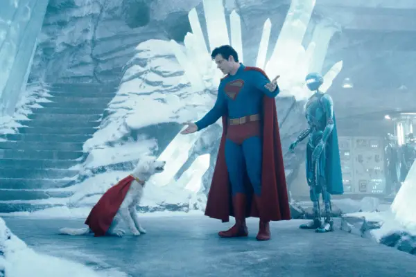 Superman (David Corenswet) is dressed in his full superman outfit (blue latex body suit, red underwear, yellow belt, black boots and red cape) is seen talking to his dog, Krypto – a white terrier mutt with a red cape on – in the Fortress of Solitude – a cave made entirely of ice. A silver robot is seen behind Superman.