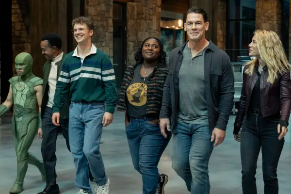 (L-R) Nhut Le, Tim Meadows, Freddie Stroma, Danielle Brooks, John Cena and Jennifer Holland star in Peacemaker; in this scene, their characters are walking shoulder-to-shoulder smiling