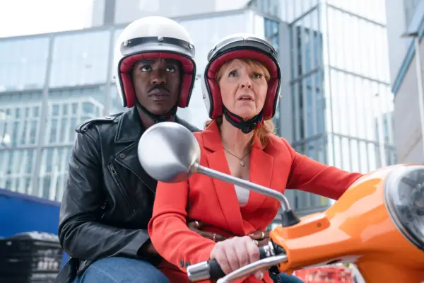 Ncuti Gatwa as The Doctor and Bonnie Langford as Mel in Doctor Who. She is riding a moped and he is sat on the back