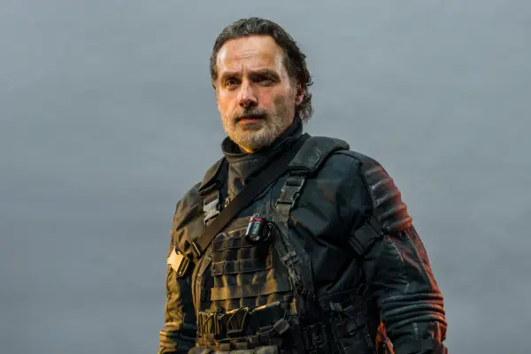 Andrew Lincoln as Rick Grimes in The Walking Dead: The Ones Who Live, wearing dark combat uniform and standing against a grey sky backdrop.
