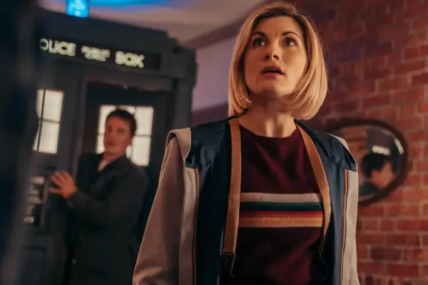 Jodie Whittaker as The Doctor in Doctor Who wearing a purple jumper and blue-grey jacket, with a man and the TARDIS blurred in the background