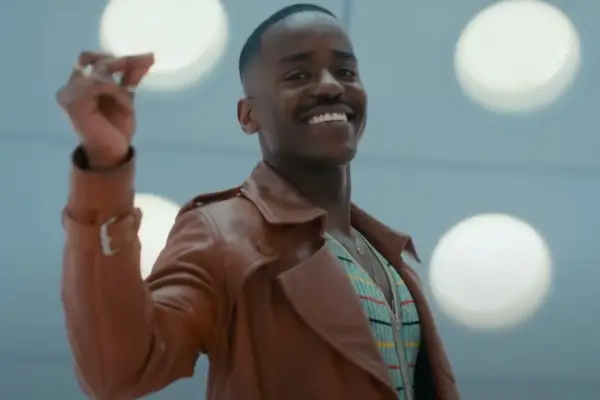 Ncuti Gatwa in Doctor Who – he is wearing a brown leather jacket and a turquoise shirt, he is smiling and clicking his fingers and is stood inside the TARDIS