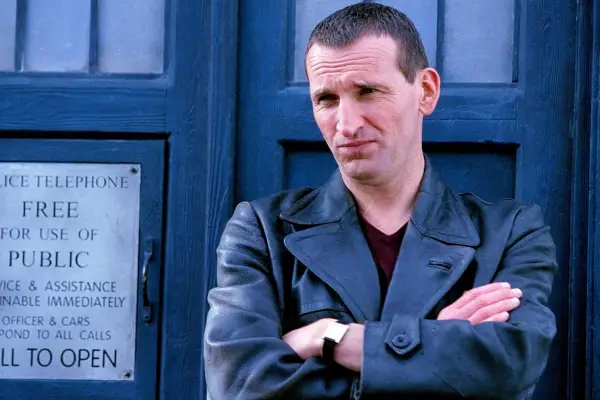 Christopher Eccleston wearing a leather jacket, arms folded, in front of the TARDIS