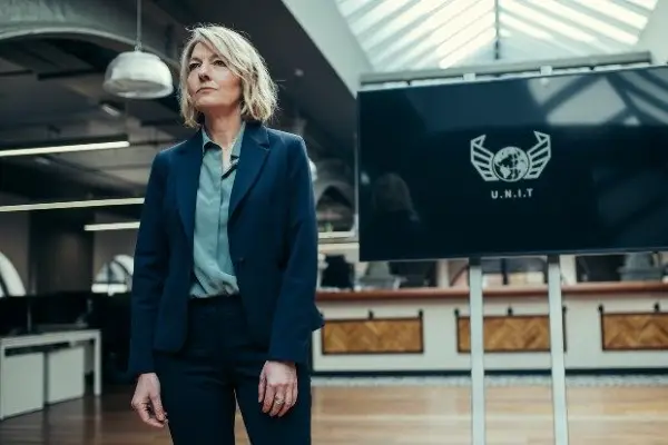 Jemma Redgrave as Kate Lethbridge-Stewart in Doctor Who season 13 standing wearing a suit, with a screen showing the UNIT logo in the background