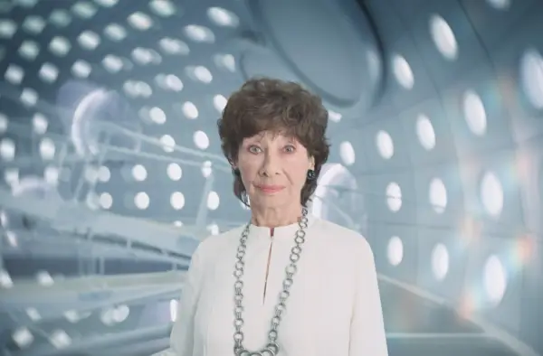 Carole Ann Ford as Susan Foreman in a white outfit and silver necklace staring at the camera and standing inside the TARDIS in Doctor Who: The Interstellar Song Contest.