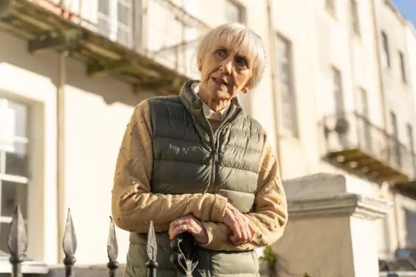 Anita Dobson as Mrs. Flood in Doctor Who. She is wearing a dark coloured gilet and tanned shirt, with her hands crossed as she looks across at something with confusion.