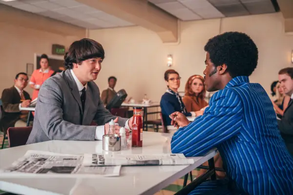 George Caple as Paul McCartney and Ncuti Gatwa as The Doctor in Doctor Who. They are sat talking in a cafe and the other patrons are looking at them.