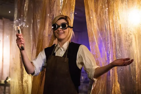 Jodie Whittaker as the Thirteenth Doctor holding a sonic screwdriver. She is wearing large goggles and is smiling.