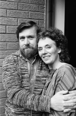 Bearded man in striped sweater hugging a woman against a brick wall.
