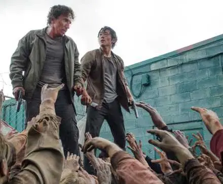 Steven Yeun as Glenn and Michael Traynor as Nicholas stand on a bin as zombies reach up in The Walking Dead.