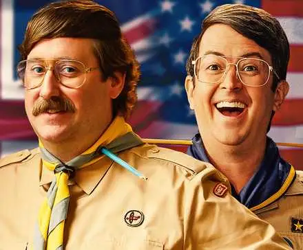 Jim Howick and Richie Moriarty both dressed in troop leader outfits in front of a US flag that