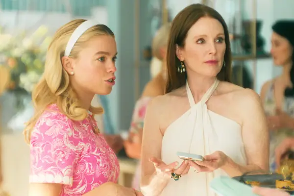 Milly Alcock as Simone, wearing a pink floral dress and white headband stood next to Julianne Moore as Michaela, wearing a white halterneck dress and holding a blue biscuit. They are attending some sort of gathering or function. A couple of other women can be seen talking in the background. There are flowers and there is food