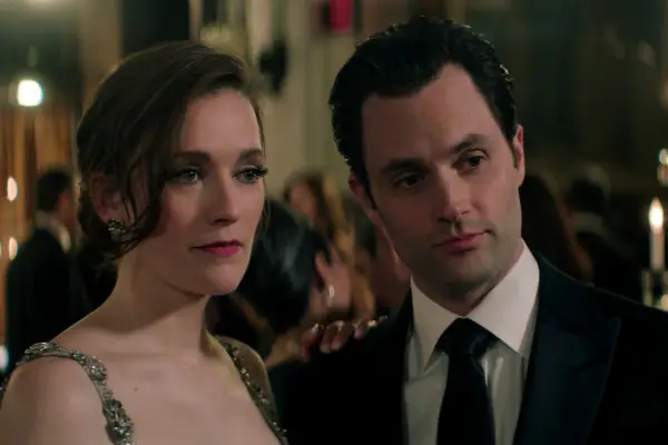 Charlotte Ritchie as Kate Lockwood, Penn Badgley as Joe Goldberg in You season 5 attending a gala and standing close together, listening to someone talk while dressed in formal attire.