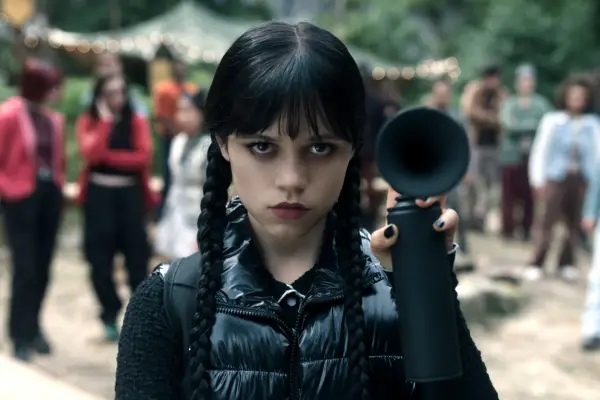Jenna Ortega as Wednesday Addams in Wednesday holding up a black horn as she stands outside amongst a crowd of people with a serious expression on her face.