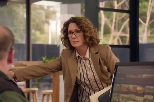 Kitty Flanagan as Helen Tudor-Fisk in the Australian sitcom Fisk; she is speaking to someone at their desk in an office setting, while reaching over to hold onto something
