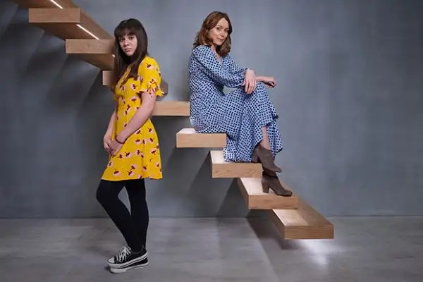 Finding Alice stars Isabella Pappas and Keeley Hawes pose next to a staircase in a grey, modern room with a bare grey wall